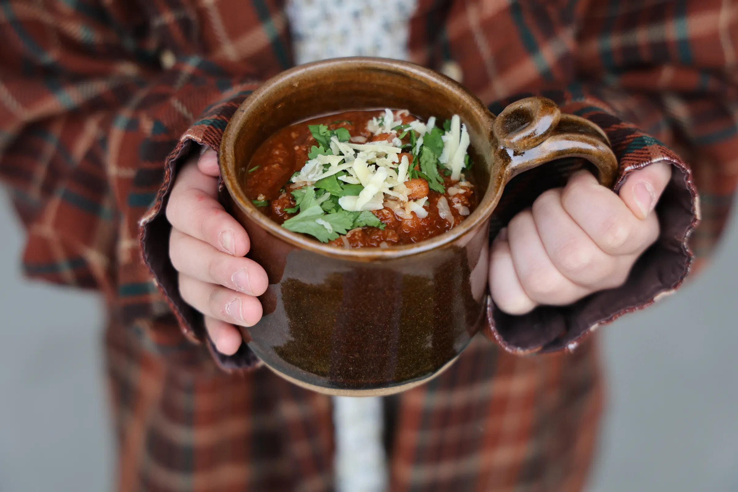 A child's hands in a flannel shirt holding out a ceramic mug of chili topped with shredded cheese and cilantro
